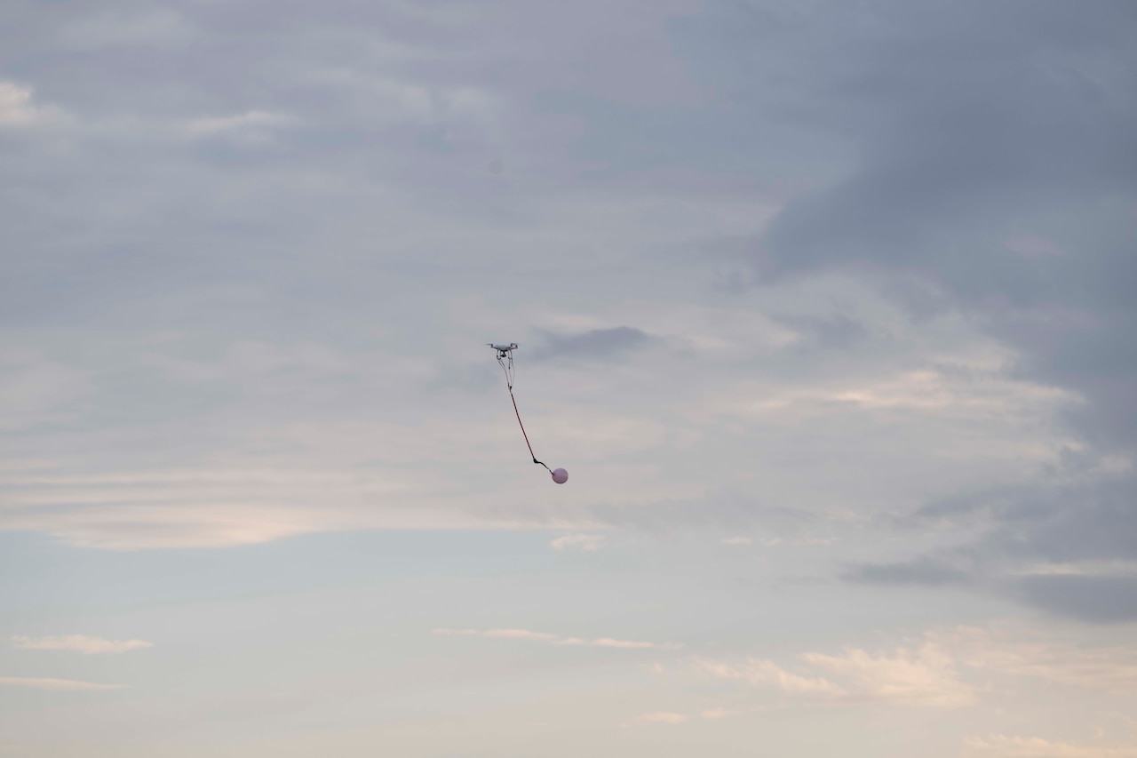 A small drone with a balloon attached flies in a blue sky with clouds.