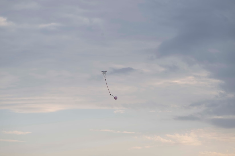 A small drone with a balloon attached flies in a blue sky with clouds.