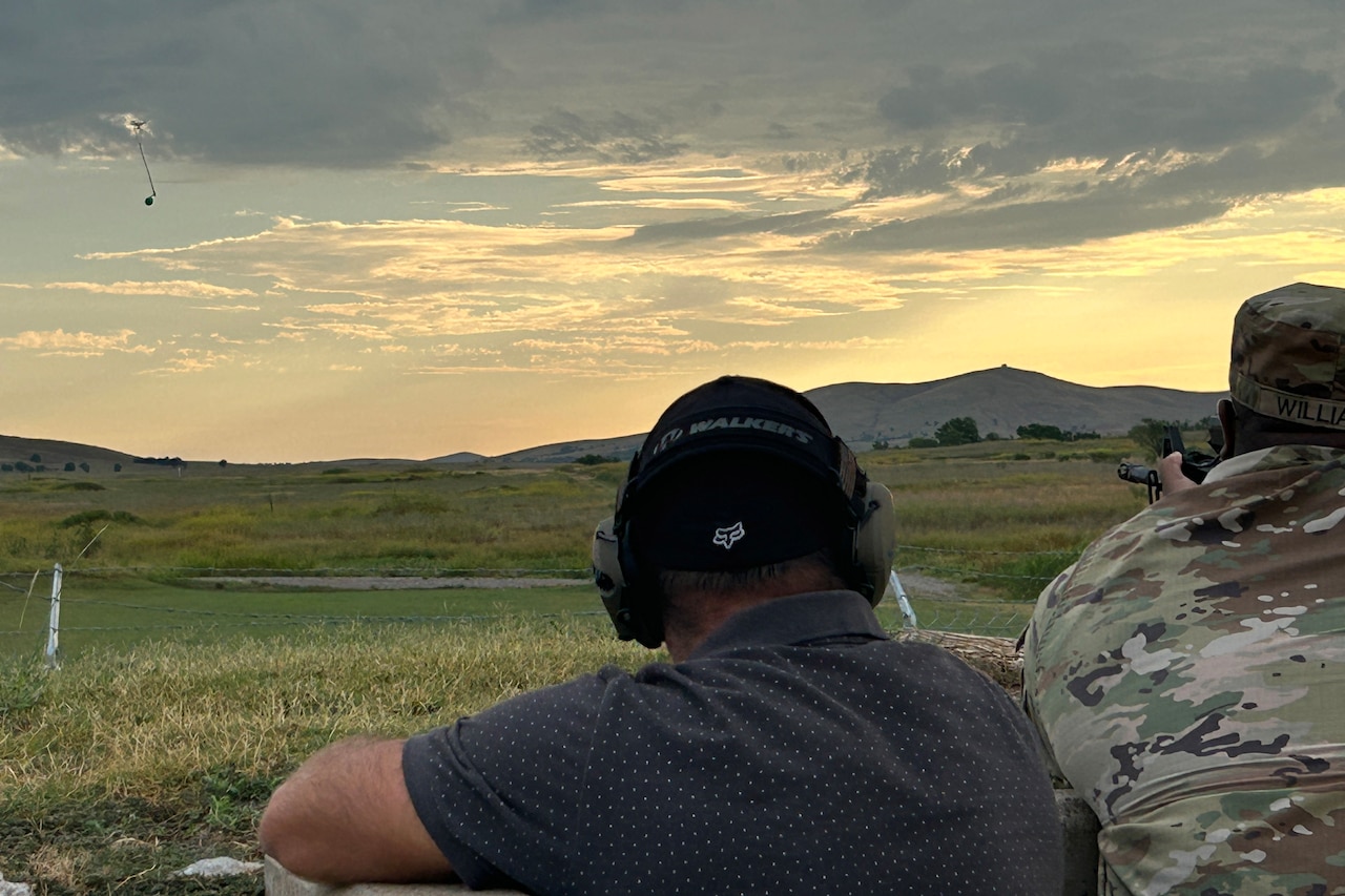 A man wearing a camouflage military uniform holds a small weapon aimed at a drone with a balloon attached. A man wearing casual attire stands beside him in a small outcrop.