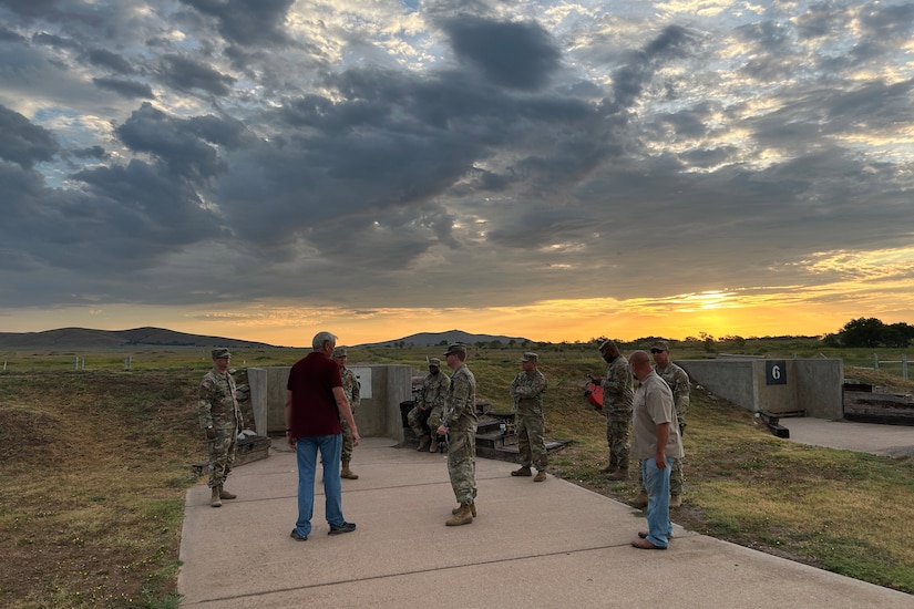 Nine men, some wearing camouflage military uniforms and others in casual attire, stand on a shooting range and talk as the sun sets behind them.
