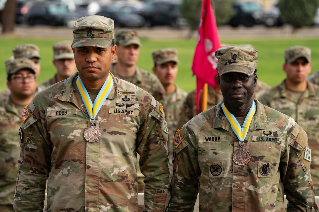 Two soldiers stand next to each other, each wearing a medal with a yellow ribbon, as others stand behind.