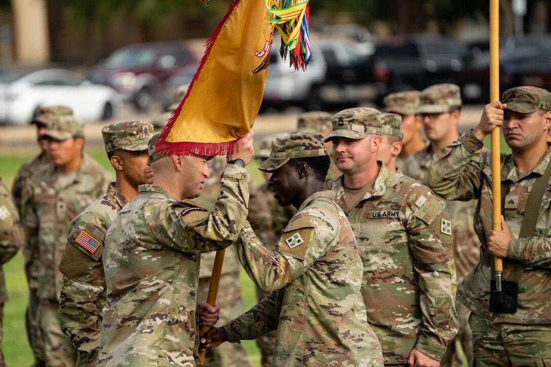 A soldier hands another soldier an orange and red flag while other soldiers watch from behind.
