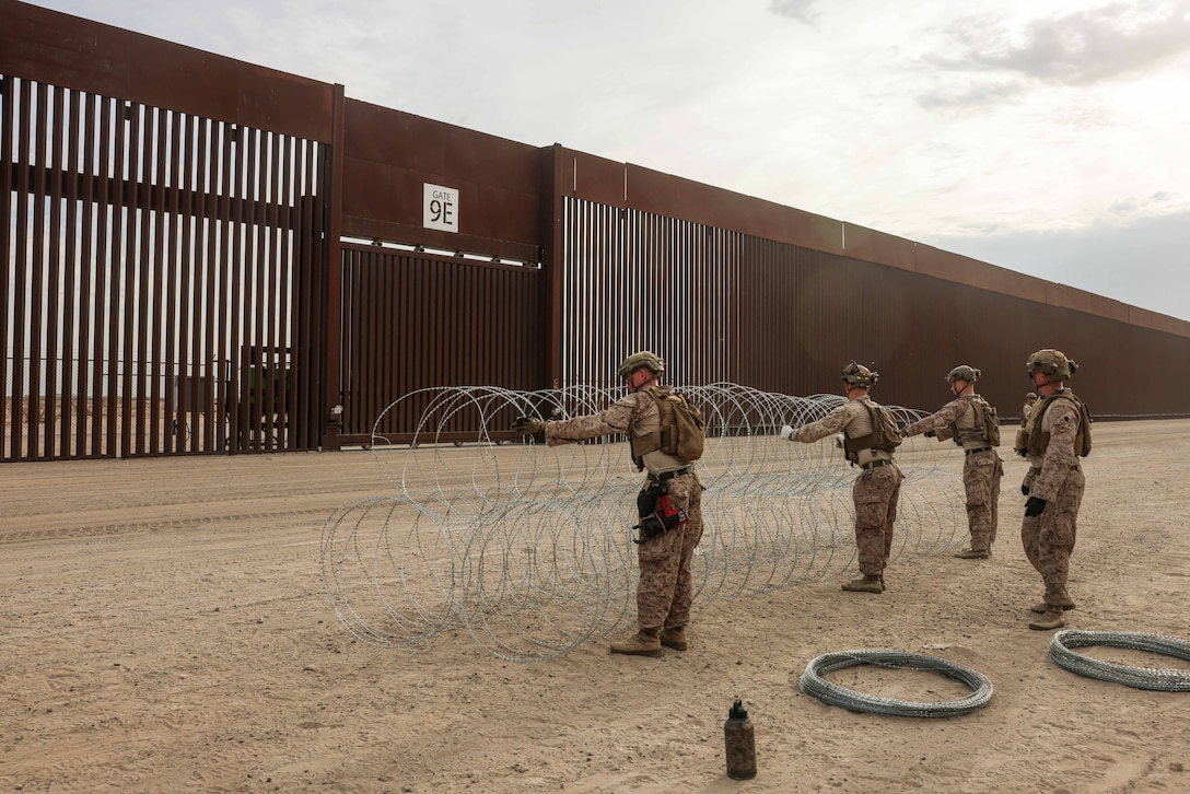 Four Marines and sailors stand on a dirt path and hold concertina wire near a tall metal wall.