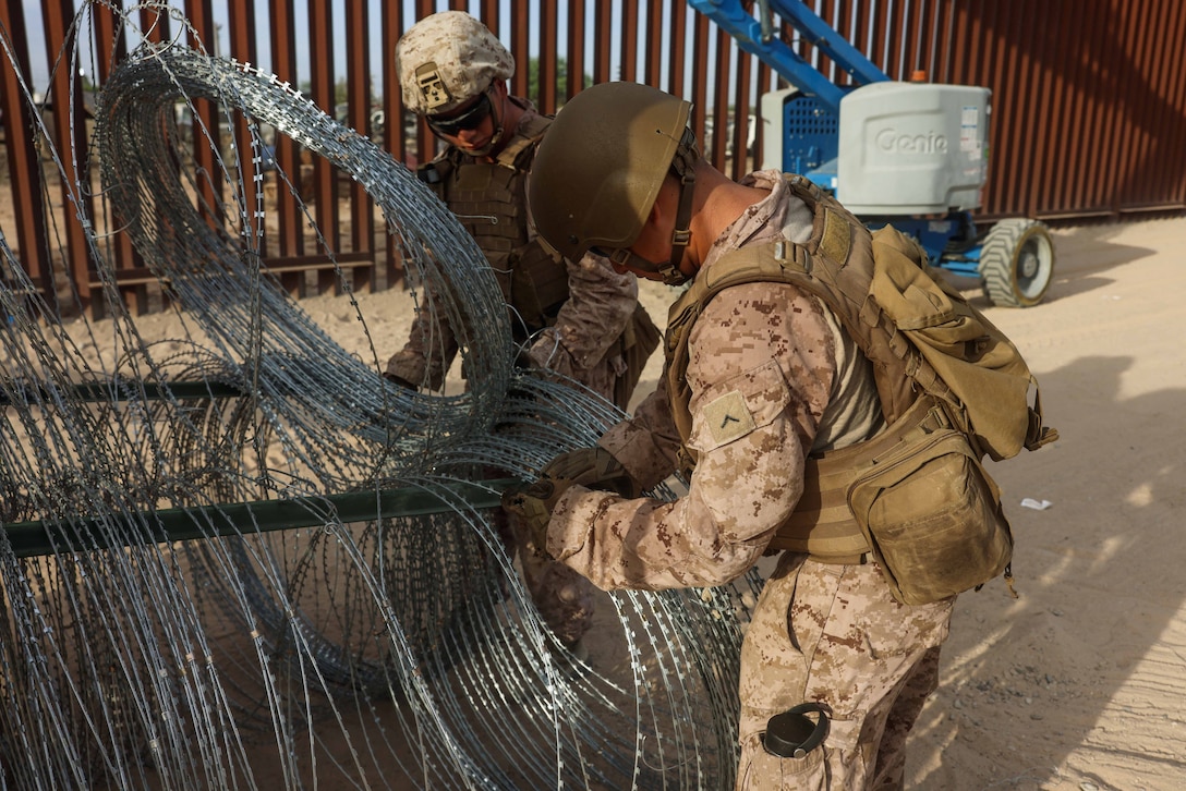 Two Marines in tactical gear hold a large coil of concertina wire next to a metal wall outside during the day.