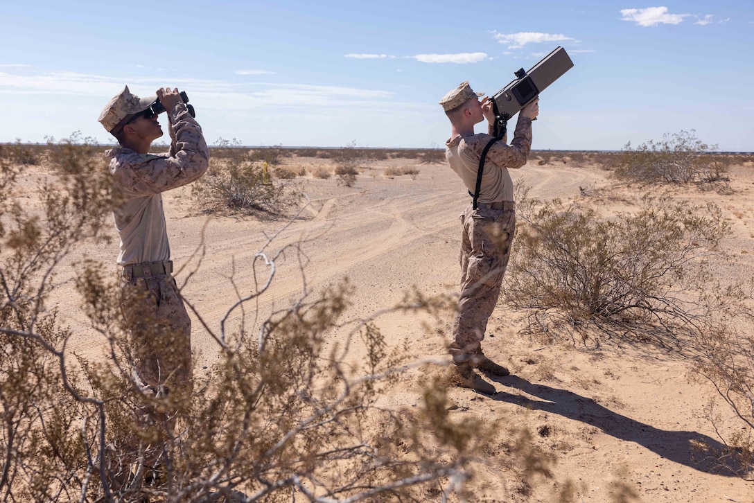 A Marine uses binoculars to look to the right while another holds up equipment in an arid setting under a blue sky.