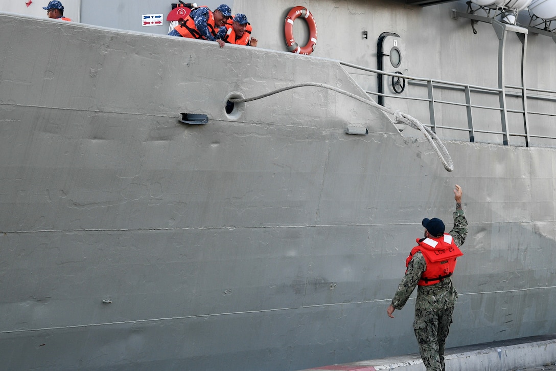 A service member releases a line as two service members watch from a ship.