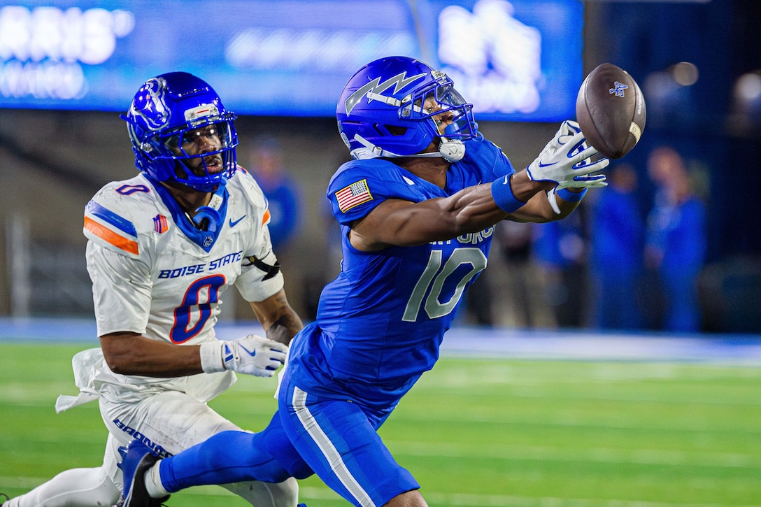 An Air Force football player reaches to catch a football during a game as an opposing player runs to catch them against a blurry background.