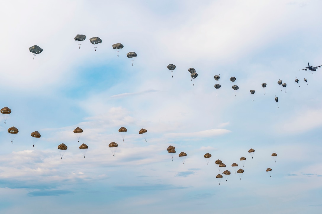 Dozens of paratroopers from allied nations descend with parachutes in a blue sky behind an aircraft.