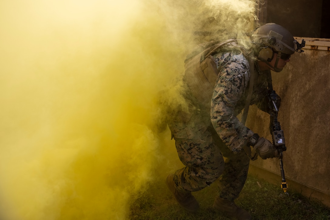 A Marine in tactical gear holds a weapon and crouches as yellow smoke billows behind.