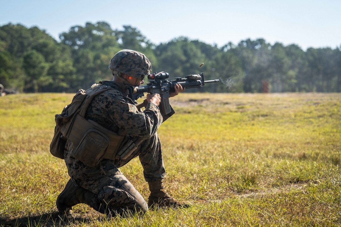 A Marine crouches in a field lined by trees during the day and fires a service weapon to the right.