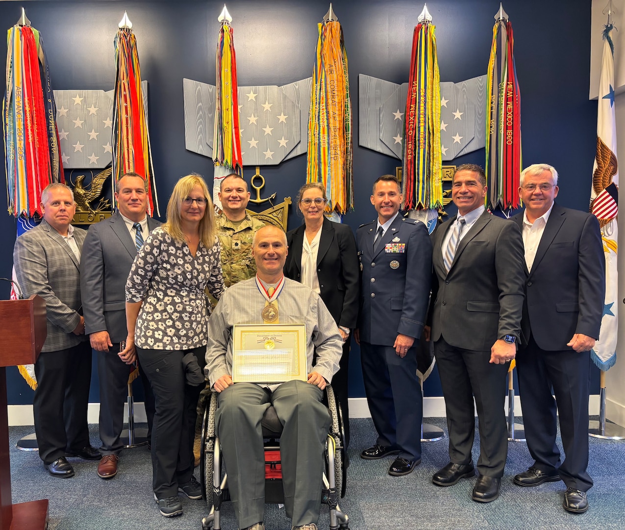 Photo of Marc Ruel in a wheelchair holding a framed award with eight people standing behind him and flags behind them.