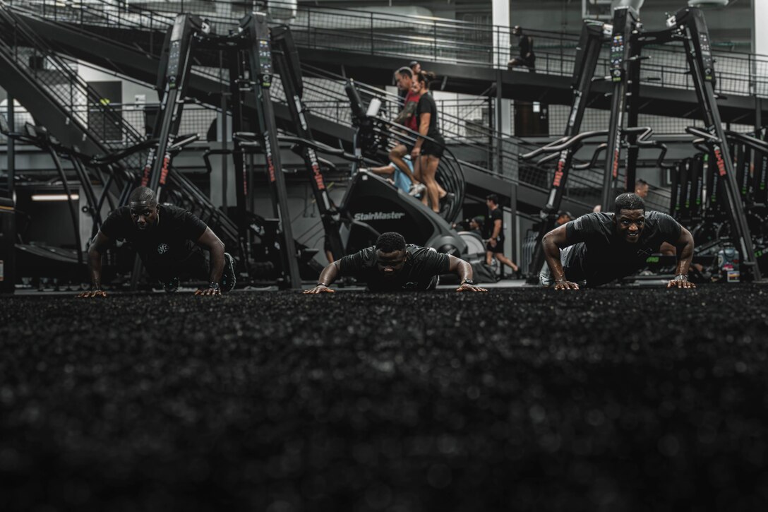 Three soldiers do pushups in a gym on a dark floor while people use gym equipment behind.