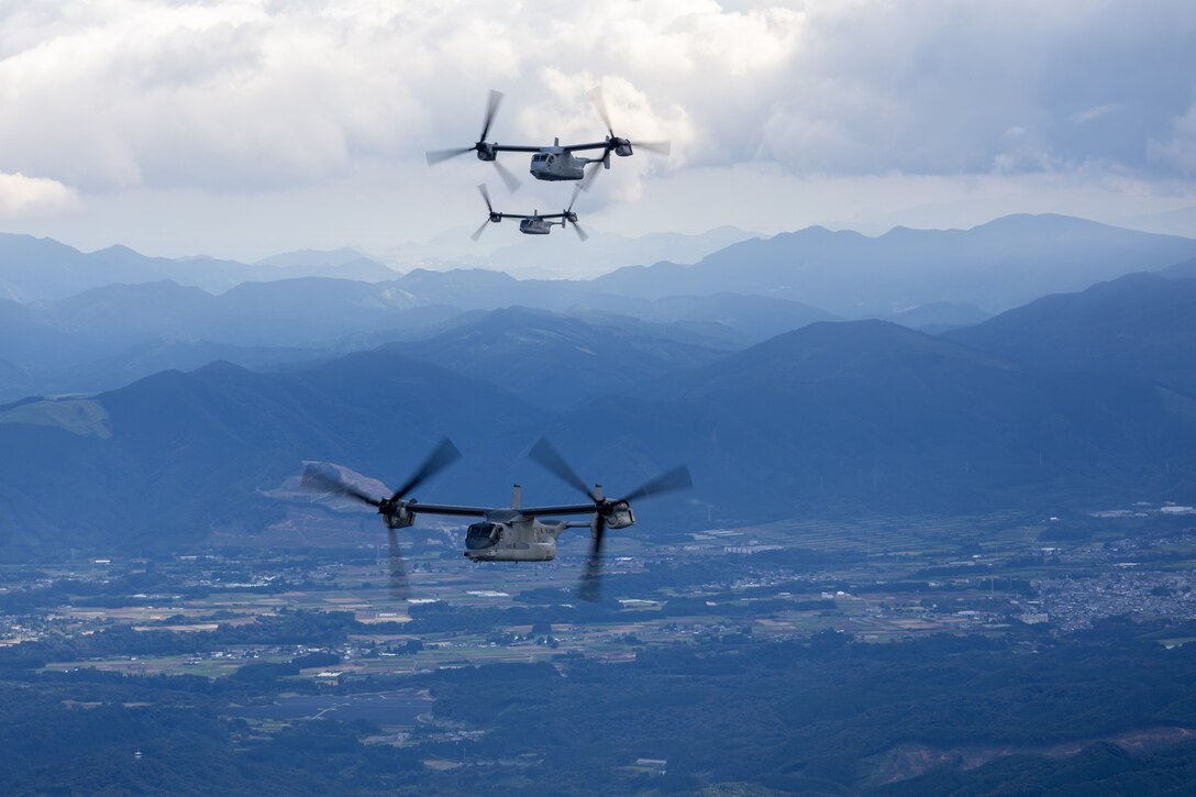 Three aircraft fly above a plain filled with fields and trees under cloudy skies with mountains in the distance.