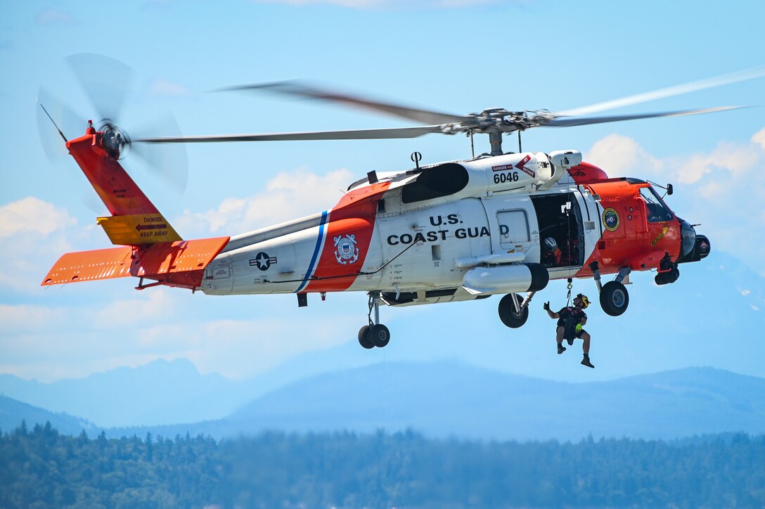 A rescue swimmer from U.S. Coast Guard Air Station Astoria dangles below an MH-60 Jayhawk helicopter as it hovers above Elliott Bay near Seattle, Washington, Aug. 1, 2023. The demonstration was part of the Parade of Ships for the annual Seafair festival. (U.S. Coast Guard photo by Petty Officer 2nd Class Steve Strohmaier)