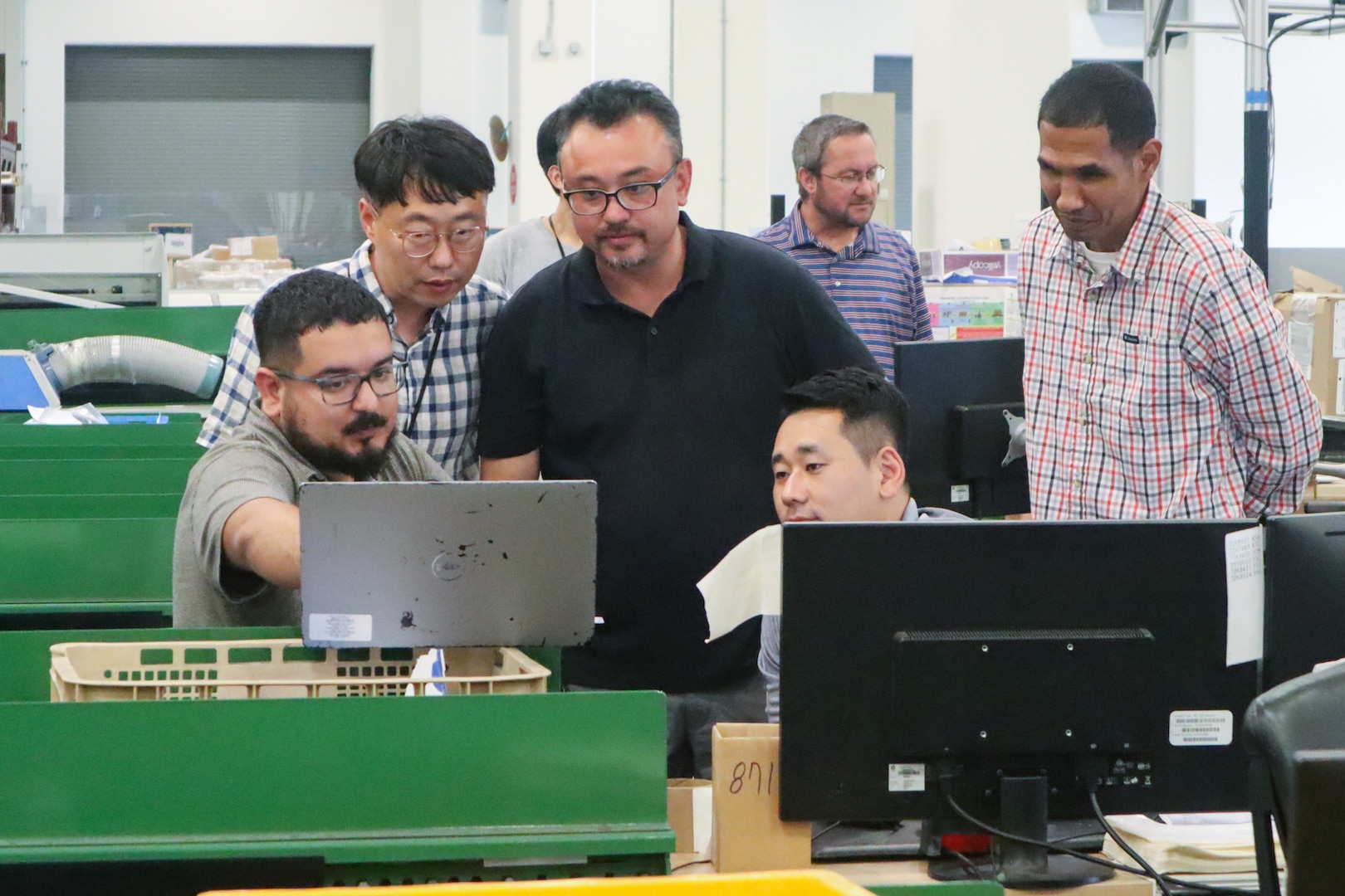 A group of people gather around a computer screen in a large warehouse. One of the people is pointing to the screen.