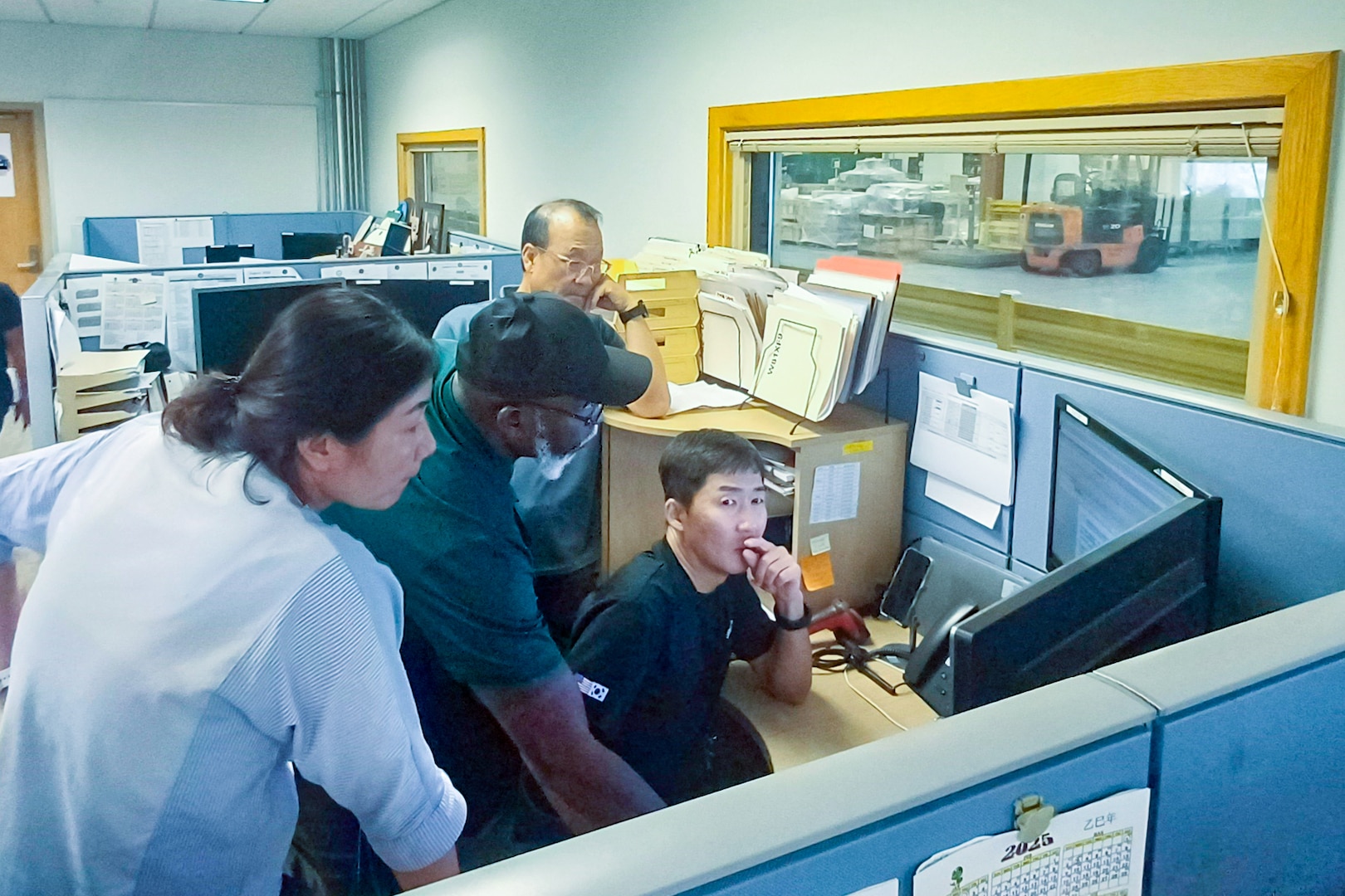 A group of people stand looking over a person working on a computer screen in a cubicle.