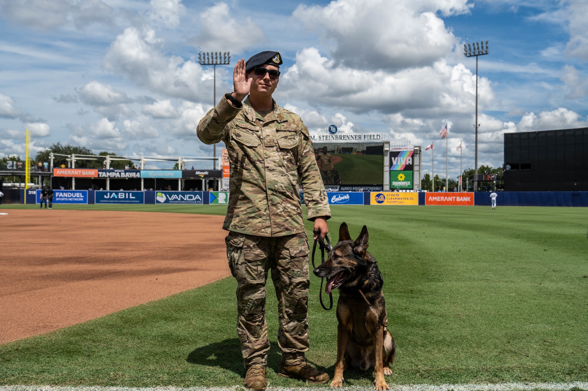 U.S. Air Force Senior Airman Jeremy Francisco, 6th Security Forces Squadron military working dog handler, and Golyko, military working dog, waves to the crowd during the Tampa Bay Rays baseball game in Tampa, Florida, Sept. 18, 2025. During the  game, Airmen assigned to the 6th ARW were recognized for their service and contributions to the community. (U.S. Air Force photo by Staff Sgt. Christopher Tam)