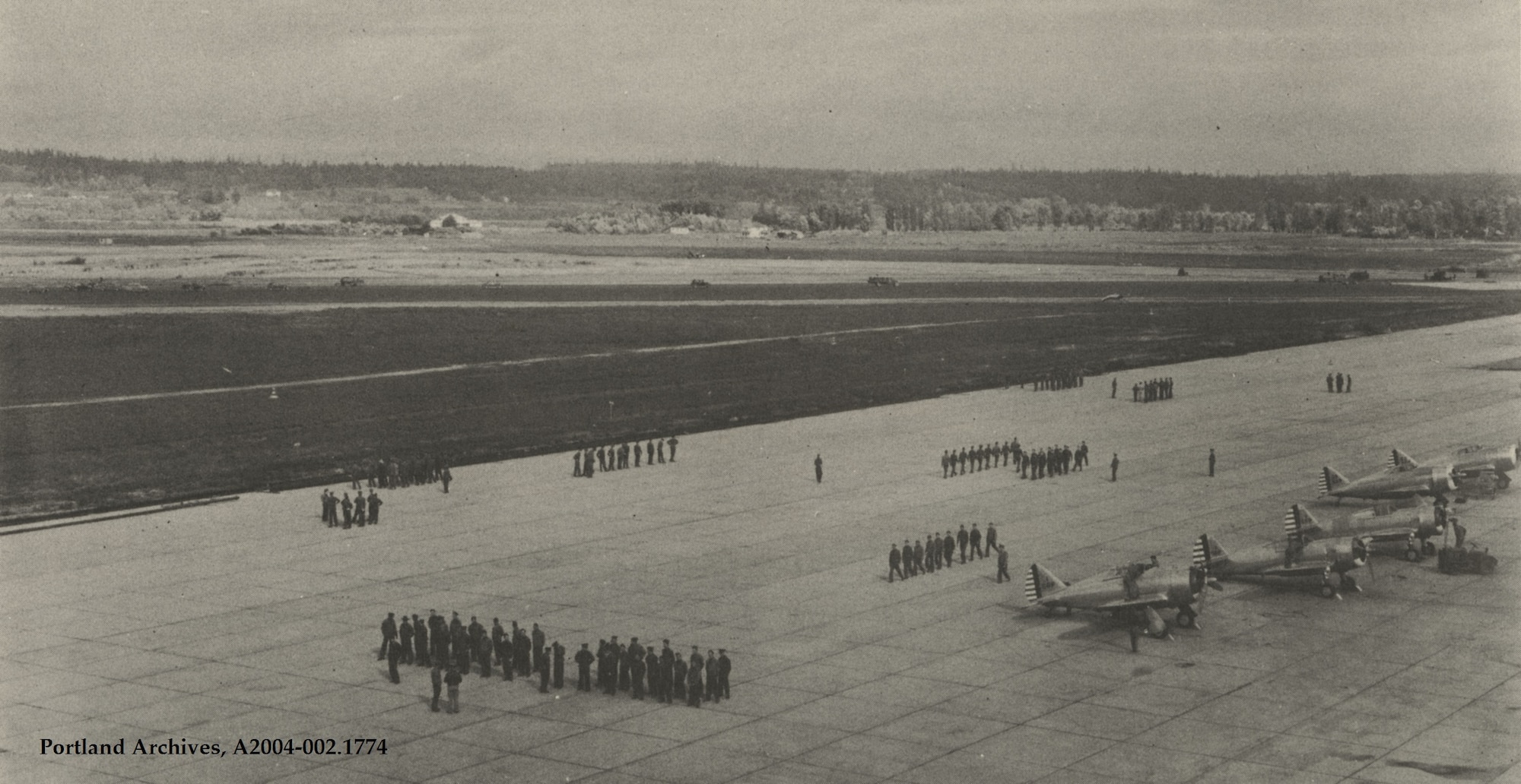 This picture of the Portland Army Air Base parking ramp is from after May, 1941 when the 55th Pursuit Group and fighter planes first arrived at Portland. It shows a mix of three Republic P-43 Lancer and two Curtiss P-36 Hawk fighter aircraft of the 55th Pursuit Group, the first flying unit based at Portland AAB. Two fighter squadrons (37th and 38th Pursuit Squadrons) flew the P-43 and another (54th Pursuit Squadron) a mix of P-43 and P-36 fighters as P-43 production worked to meet demand. The men on the ramp in various formations are perhaps drilling and marching in preparation for a military parade. Note what appears to be construction activity ongoing in the distance, as Portland’s runways and taxiways were improved to handle military aircraft carrying heavy loads.  As war neared in late 1941, the 55th Pursuit Group’s fighters were painted in what became the standard US Army Air Corps/Army Air Force camouflage of olive drab on the upper surfaces with a neutral gray undersurface. (Photo courtesy of the City of Portland Auditor’s Office Archives)