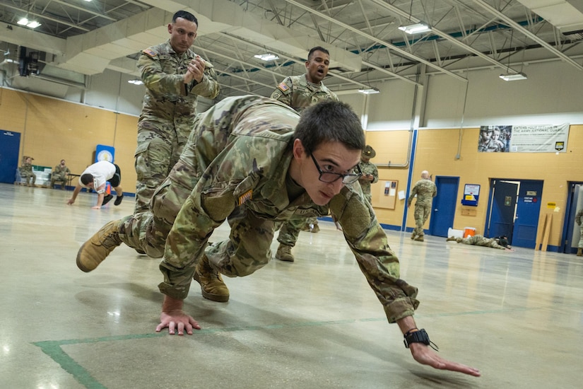 A man wearing glasses and a camouflage military uniform crawls across a gym floor as two other men, also dressed in camouflage military uniforms, yell at him.