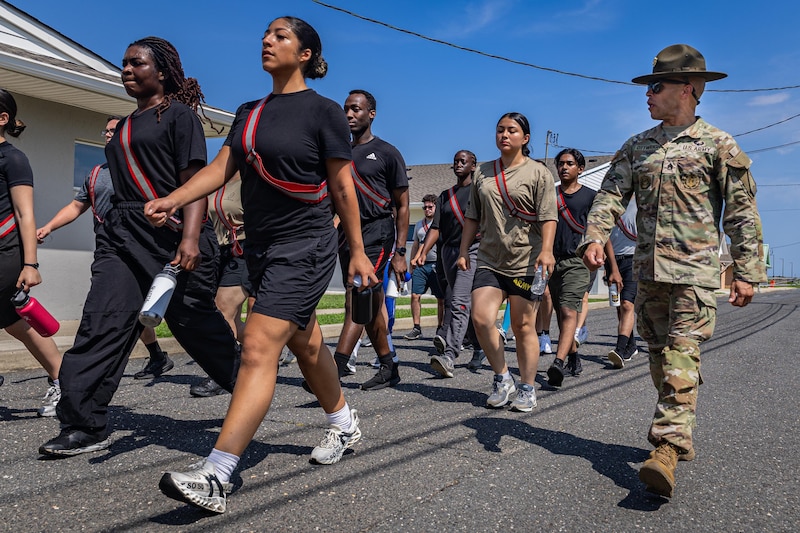 A group of men and women dressed in civilian attire marches in formation, with a man wearing a camouflage military uniform and sunglasses marching alongside the formation.