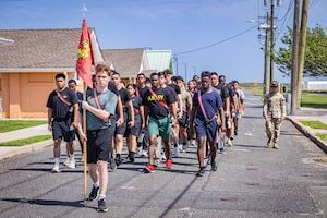 A group of men and women, all dressed in civilian attire, march in formation as a man in civilian attire leads the group, carrying a flag attached to a pole. A man wearing a camouflage military uniform marches to the right of the formation.