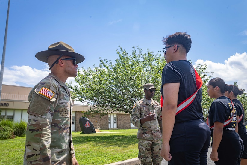 A man wearing a camouflage military uniform and sunglasses stares at a man dressed in civilian attire while standing in formation. In the background, another man, wearing a camouflage military uniform, gestures to two women in civilian attire who are also standing in formation.