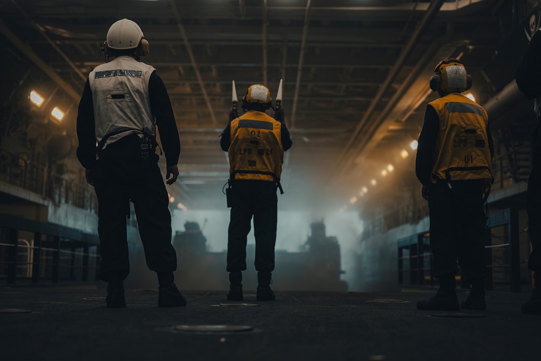 Three sailors, one holding batons up in each hand, stand in a dark hangar bay of a ship while directing an air-cushioned landing craft.