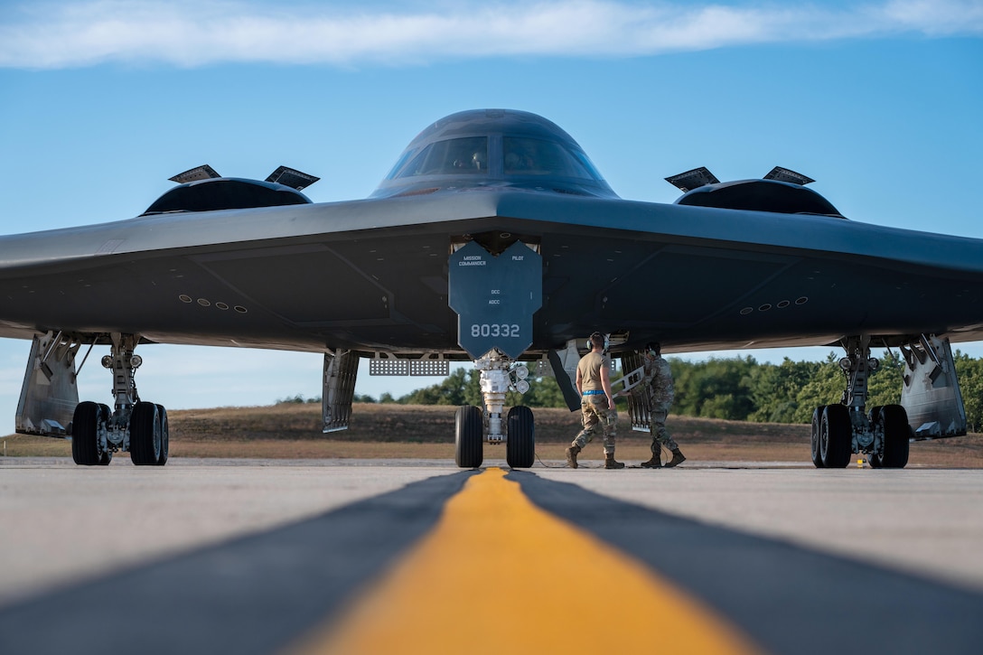 Two airmen work on an aircraft parked on a tarmac under a blue sky with greenery in the background.