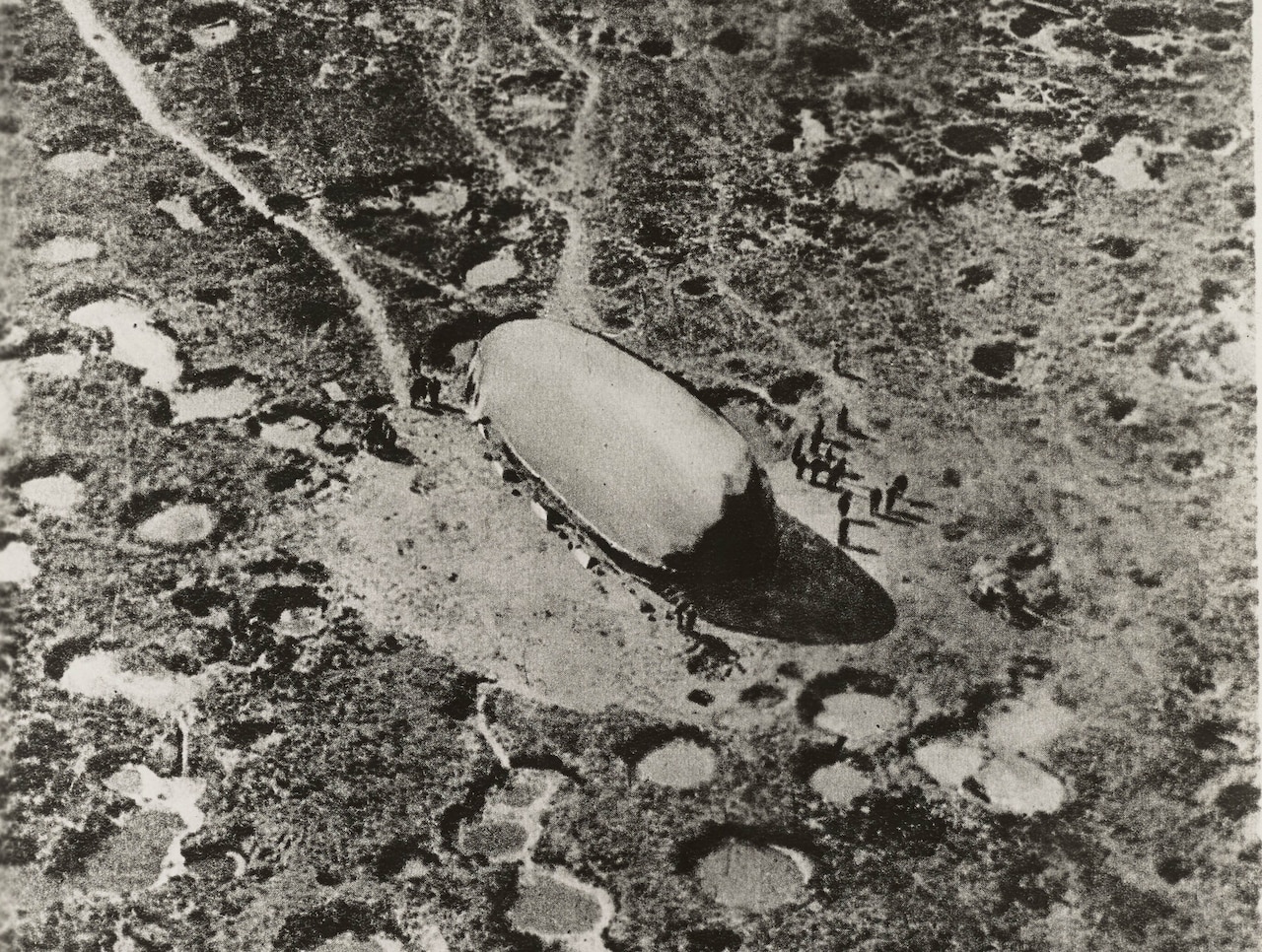A massive balloon sits on the ground with craters scattered around it. About a dozen people stand beside the balloon.