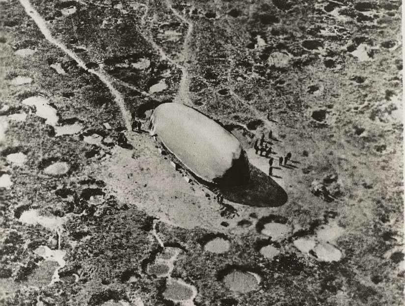 A massive balloon sits on the ground with craters scattered around it. About a dozen people stand beside the balloon.