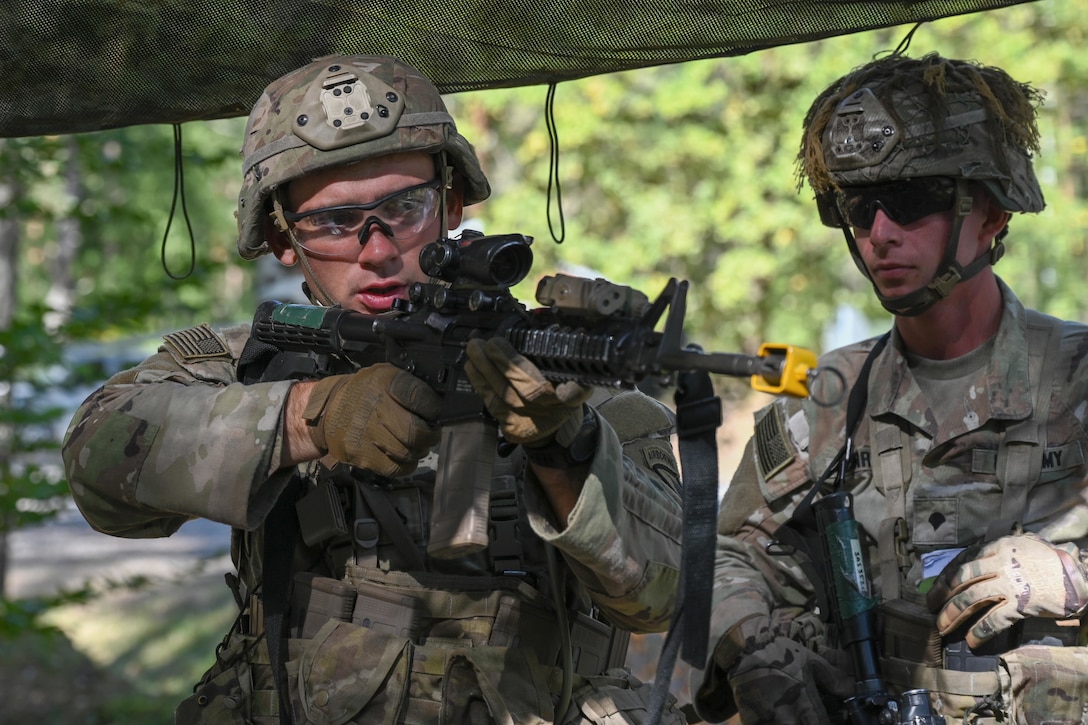 A soldier aims a weapon as a fellow soldier watches in a wooded area.