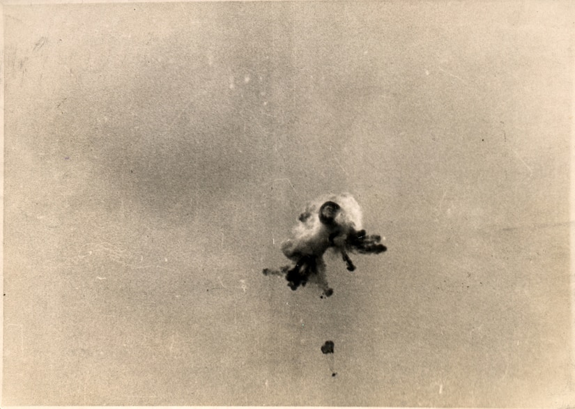 Smoke and debris hang in the sky. A small parachute can be seen below the debris.