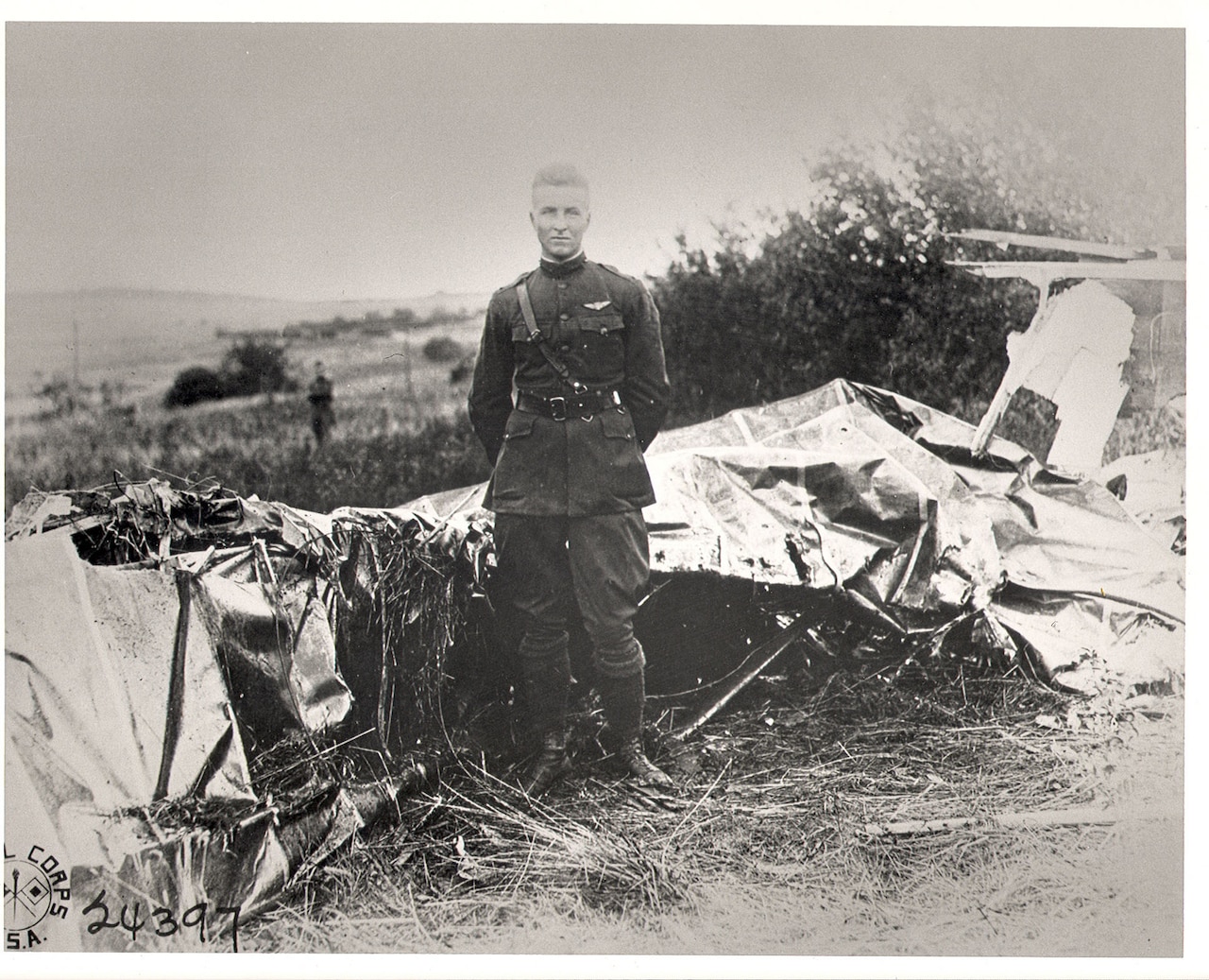 A man dressed in a military uniform stands stoically in front of a crumpled pile of metal.