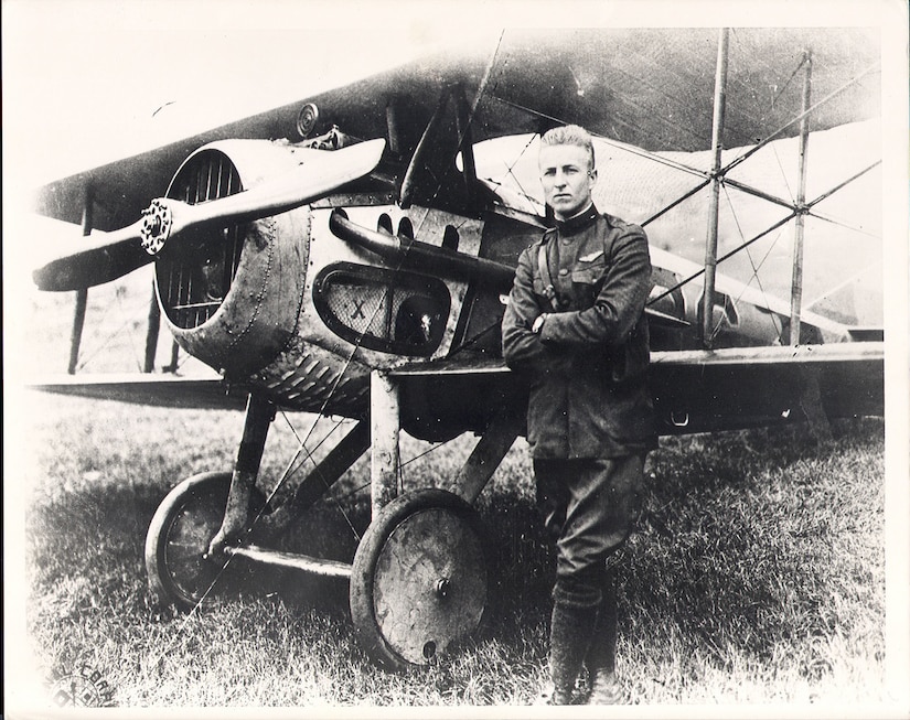 A man wearing a military uniform with his arms crossed over his chest stands in front of a propeller aircraft.