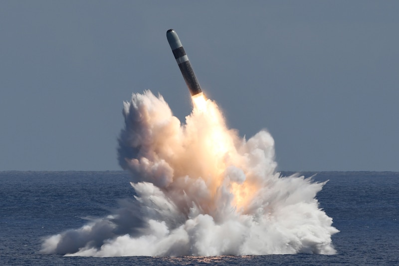A missile launches out of the ocean and leaves behind spray, smoke and fire during the day.