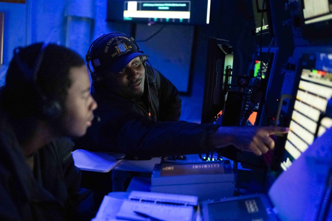A sailor sits in front of computers on a desk and points while another watches in a room illuminated by blue light.