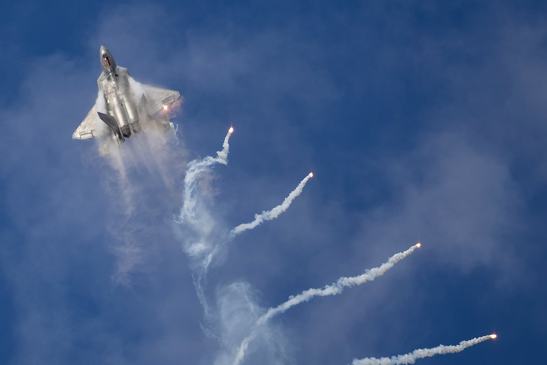 An aircraft flies above four flares and trails of smoke against a blue sky.