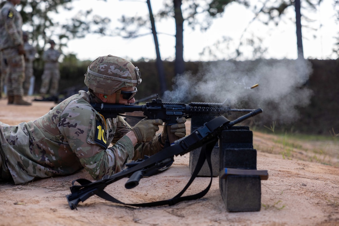 A helmeted soldier rests a service weapon on a block and shoots from the ground while outside.