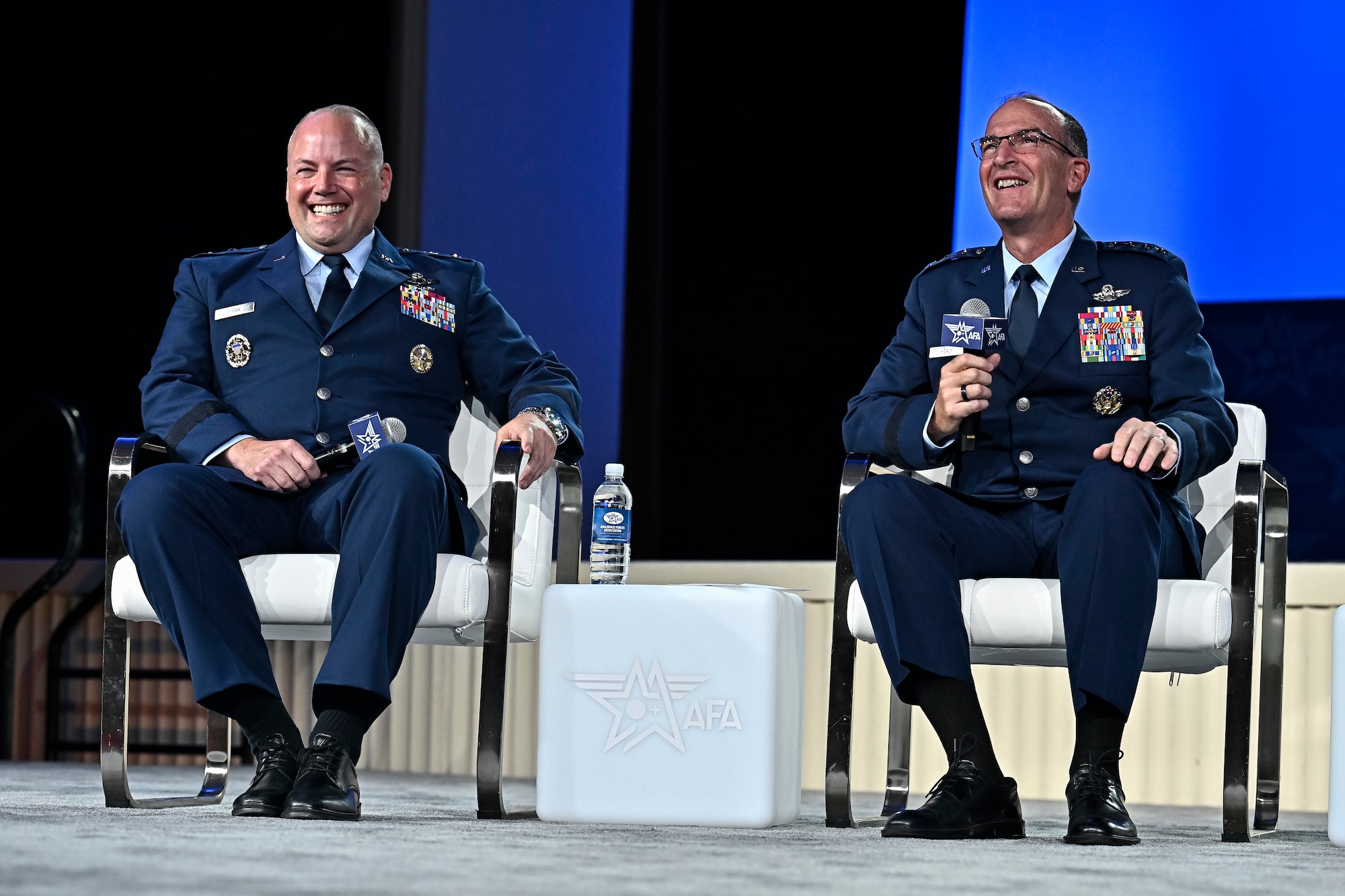Two men sit in chairs on a stage.