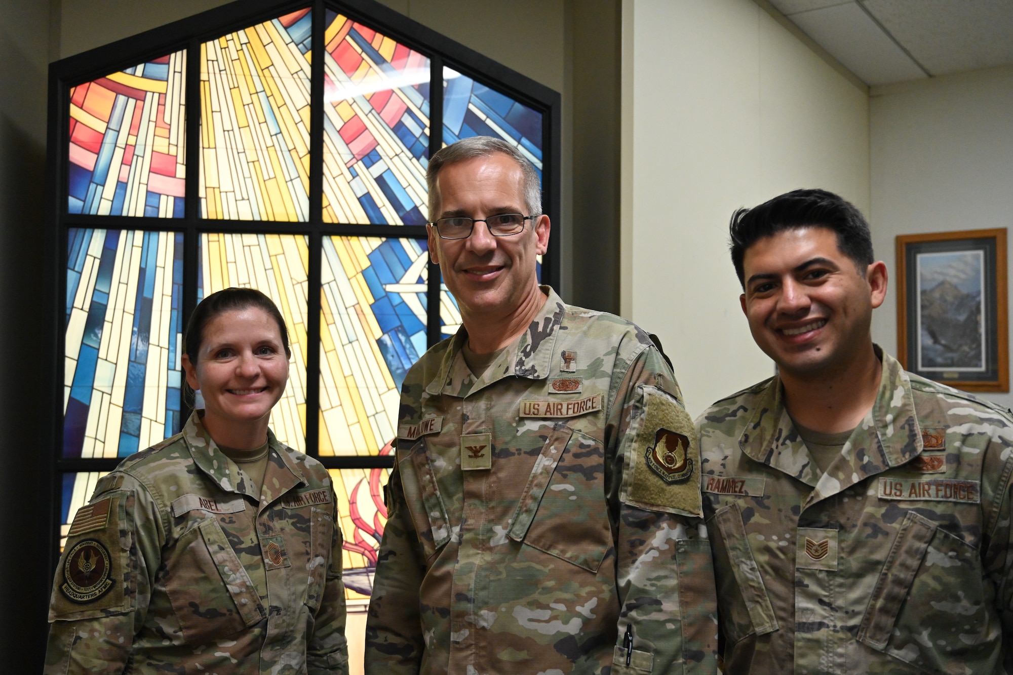 Chief Master Sgt. Jennifer Abel, Senior Enlisted Leader for Religious Affairs, Chaplain (Col.) Dale Marlowe, Air Force Materiel Command Chaplain, and Tech. Sgt. Tyler Ramirez, Readiness and Resource Support Manager, pose for a photo in the chaplain’s office at AFMC headquarters. They are wearing OCP's and standing in front of a stained-glass window.