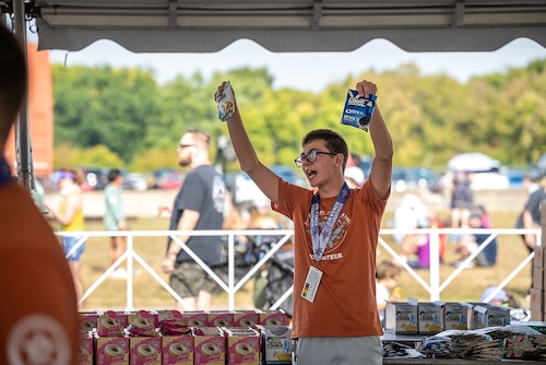 A young man, wearing glasses and orange volunteer shirt, holds up his arms high while holding different snacks