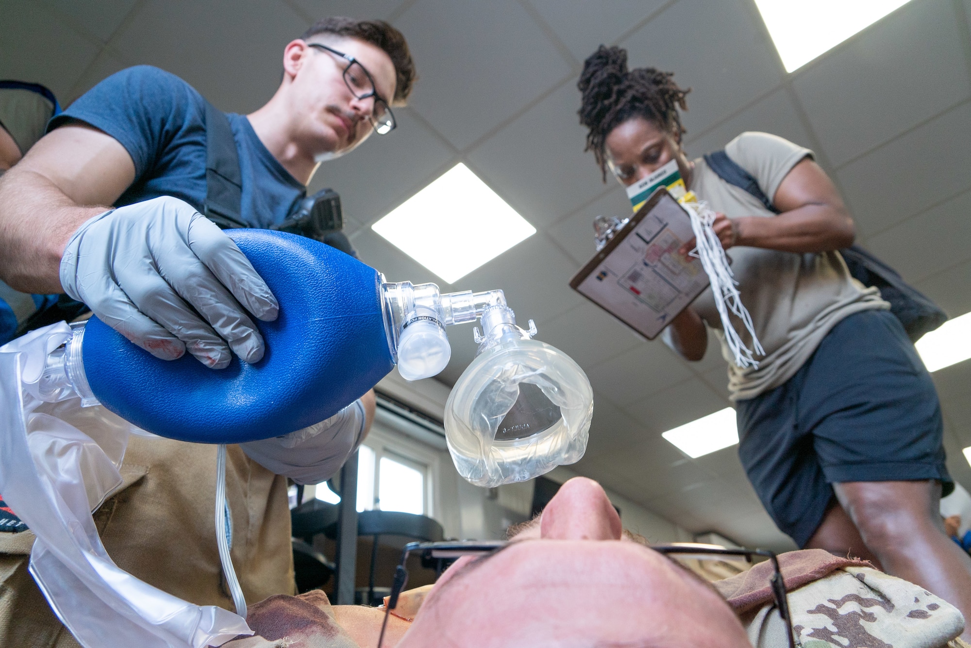 Medical Airmen on either side of frame, take care of a patient bottom of frame with the Airman on the right taking down notes on a clipboard and the Airman on the left providing simulated oxygen.