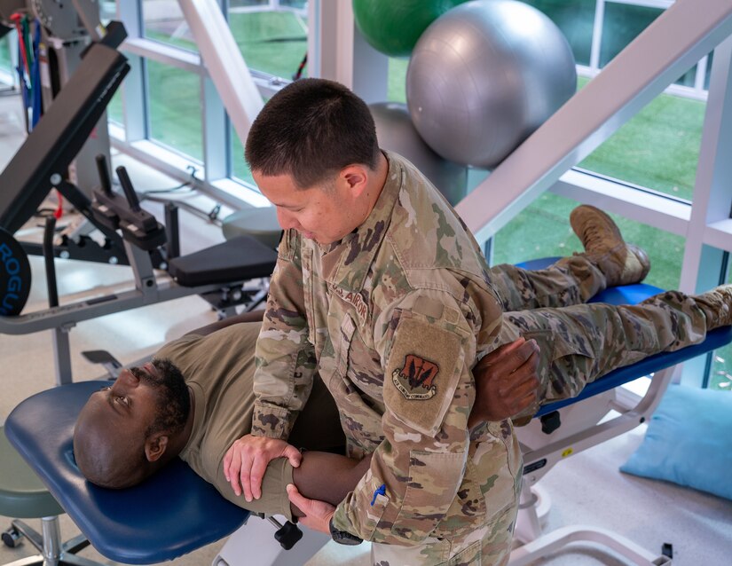 Service member presses down on another service member's arm while lying on a medical table.