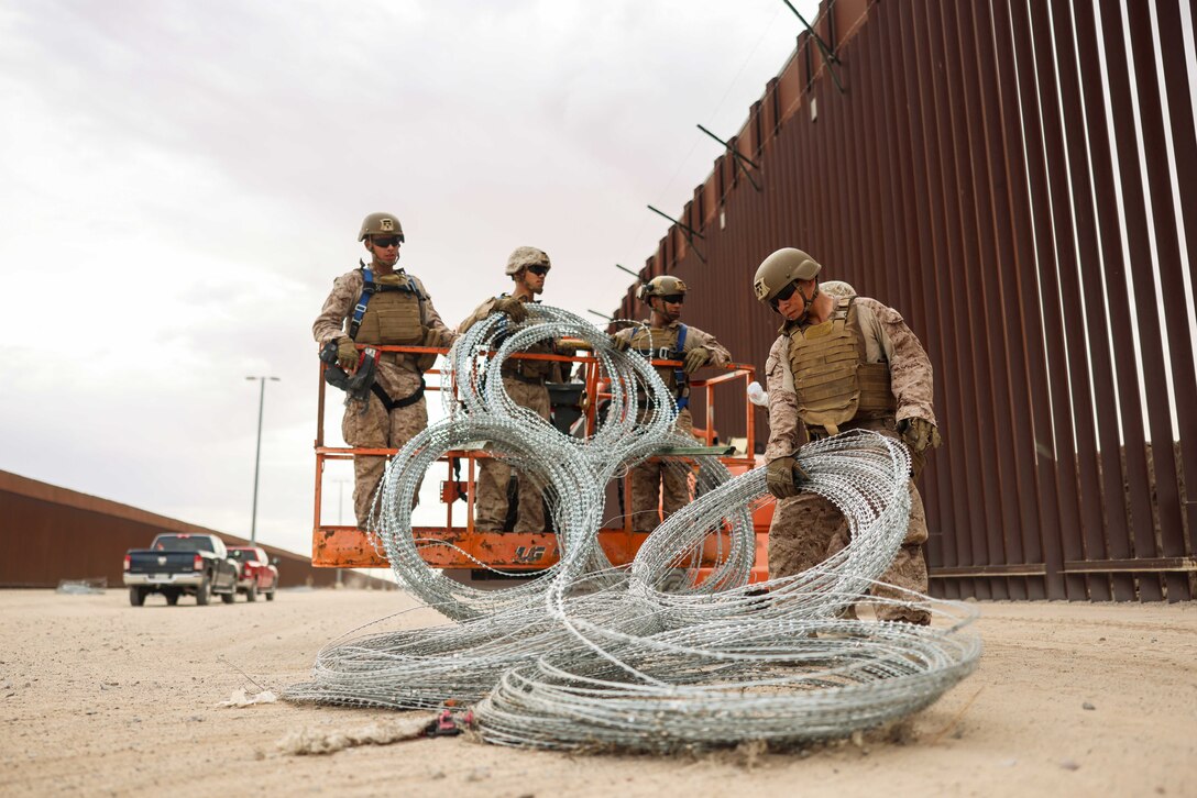 A Marine picks up coils of concertina wire as three Marines behind watch and help next to a tall metal fence.