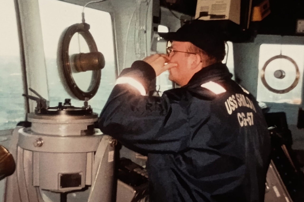 A man wearing a reflective coat and hat looks out the windows from the bridge of a military ship. The man is surrounded by nautical equipment.