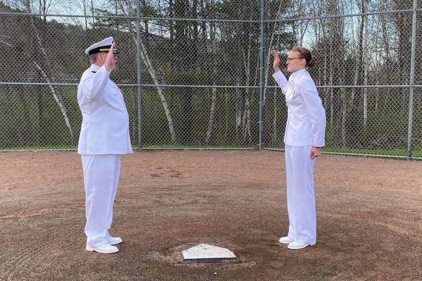 A man on the left, wearing a white military dress uniform, stands with his right hand raised as a woman in similar attire stands to his right, doing the same thing. They are both standing on opposite sides of home plate on a baseball field.