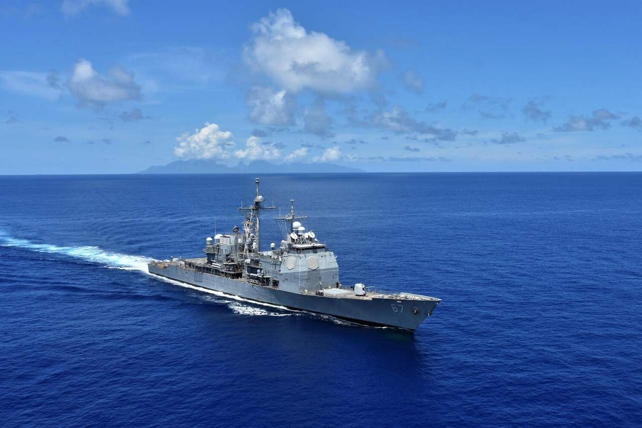 A large gray military ship sails in the ocean during a partly cloudy day.