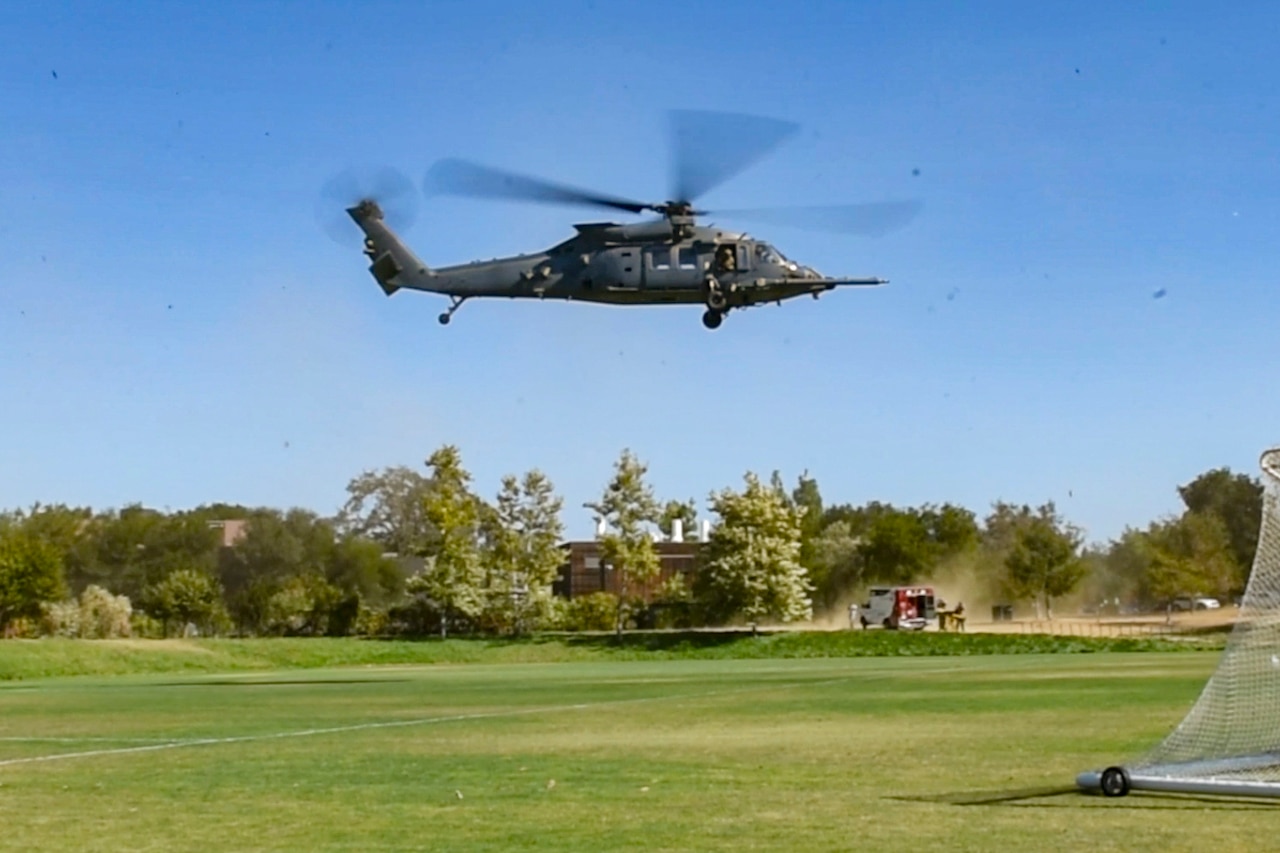 A helicopter hovers in a blue sky over a grassy field with trees in the background and a soccer goal in the foreground.