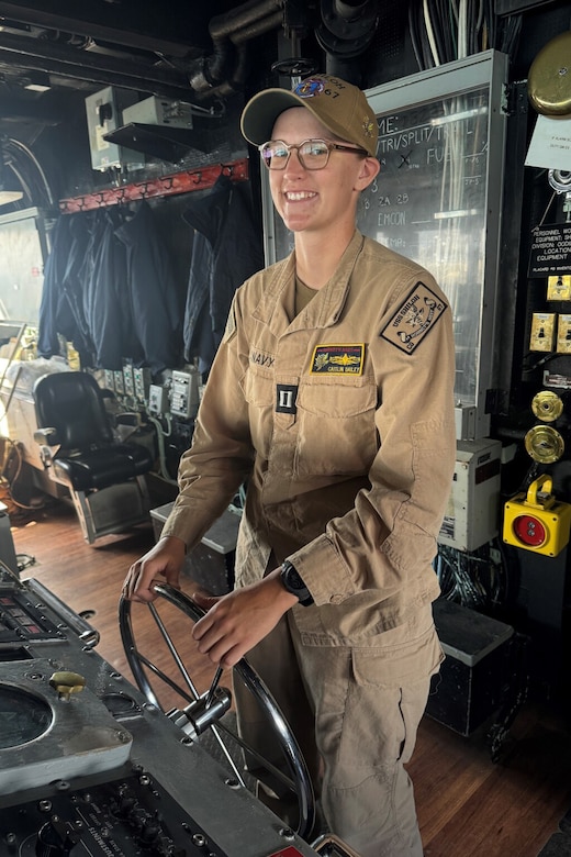 A woman wearing a khaki military uniform smiles while steering a military ship.