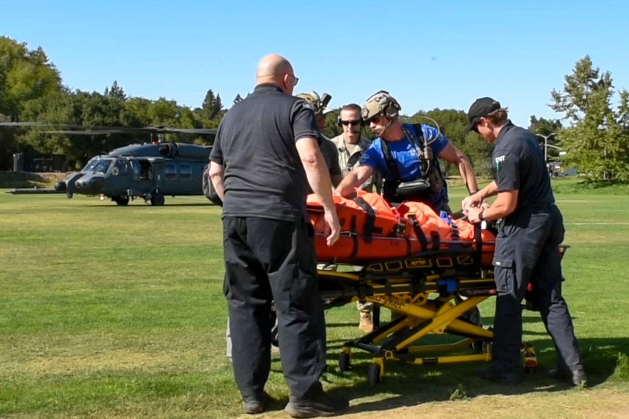Five people secure a person to a medical transport stretcher in a grassy field with a helicopter behind them.
